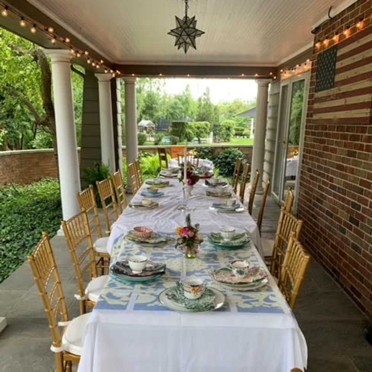 A long dining table is set for a meal on a covered patio, surrounded by greenery and decorated with dishes and floral centerpieces.