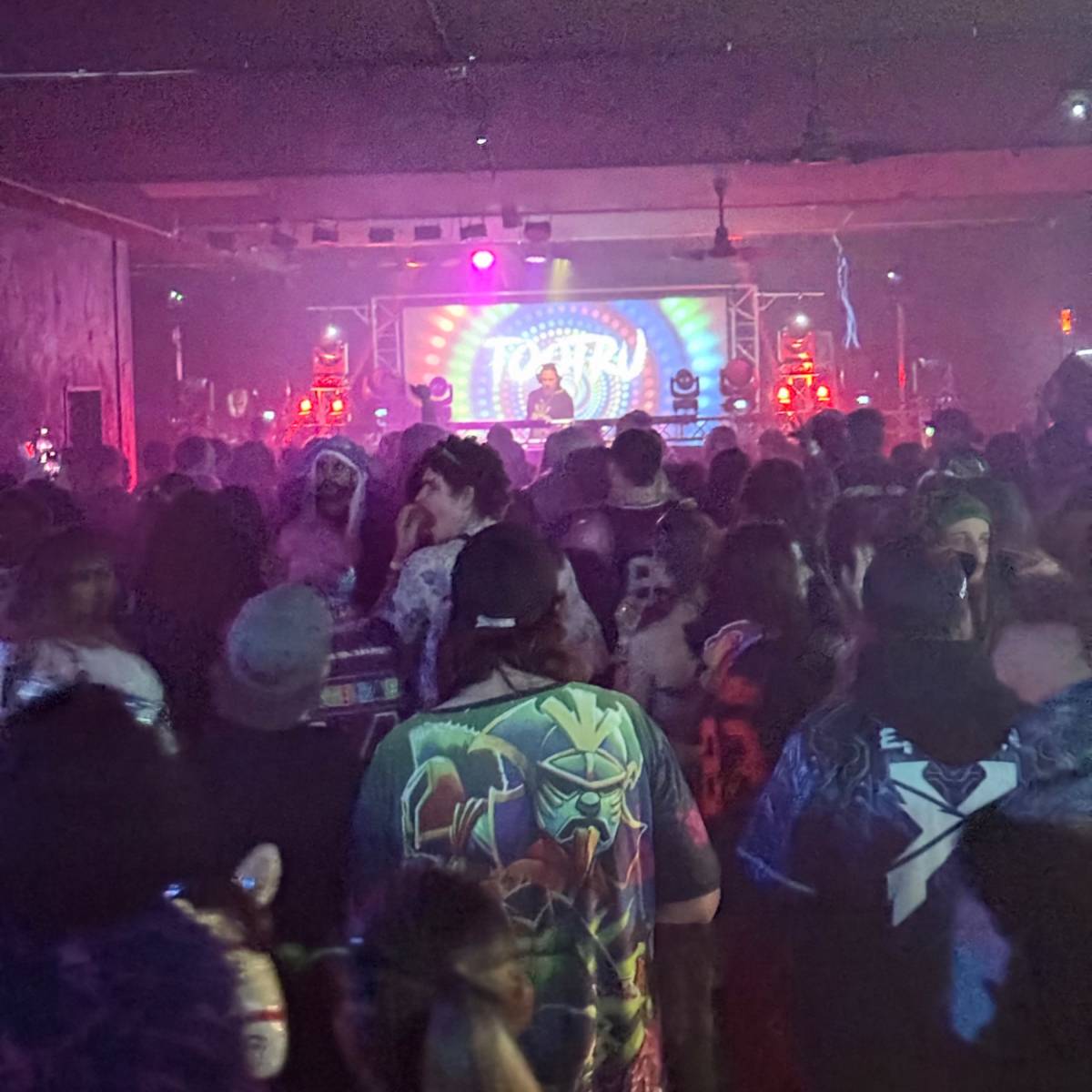 A crowded nightclub scene features a DJ performing on stage with colorful lights flashing in the background.