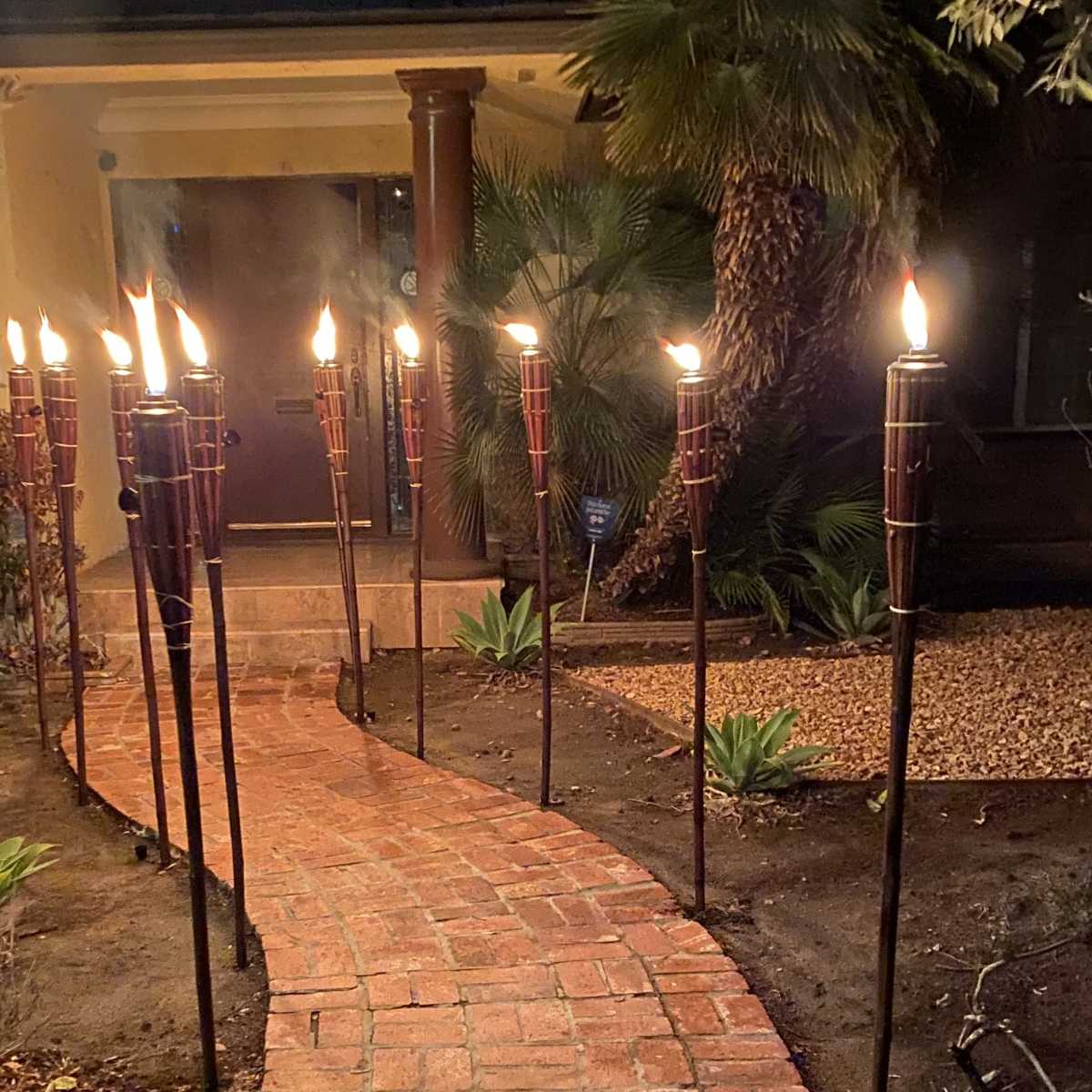 A brick pathway, lined with lit torches, leads to the entrance of a house surrounded by palm trees and landscaping.