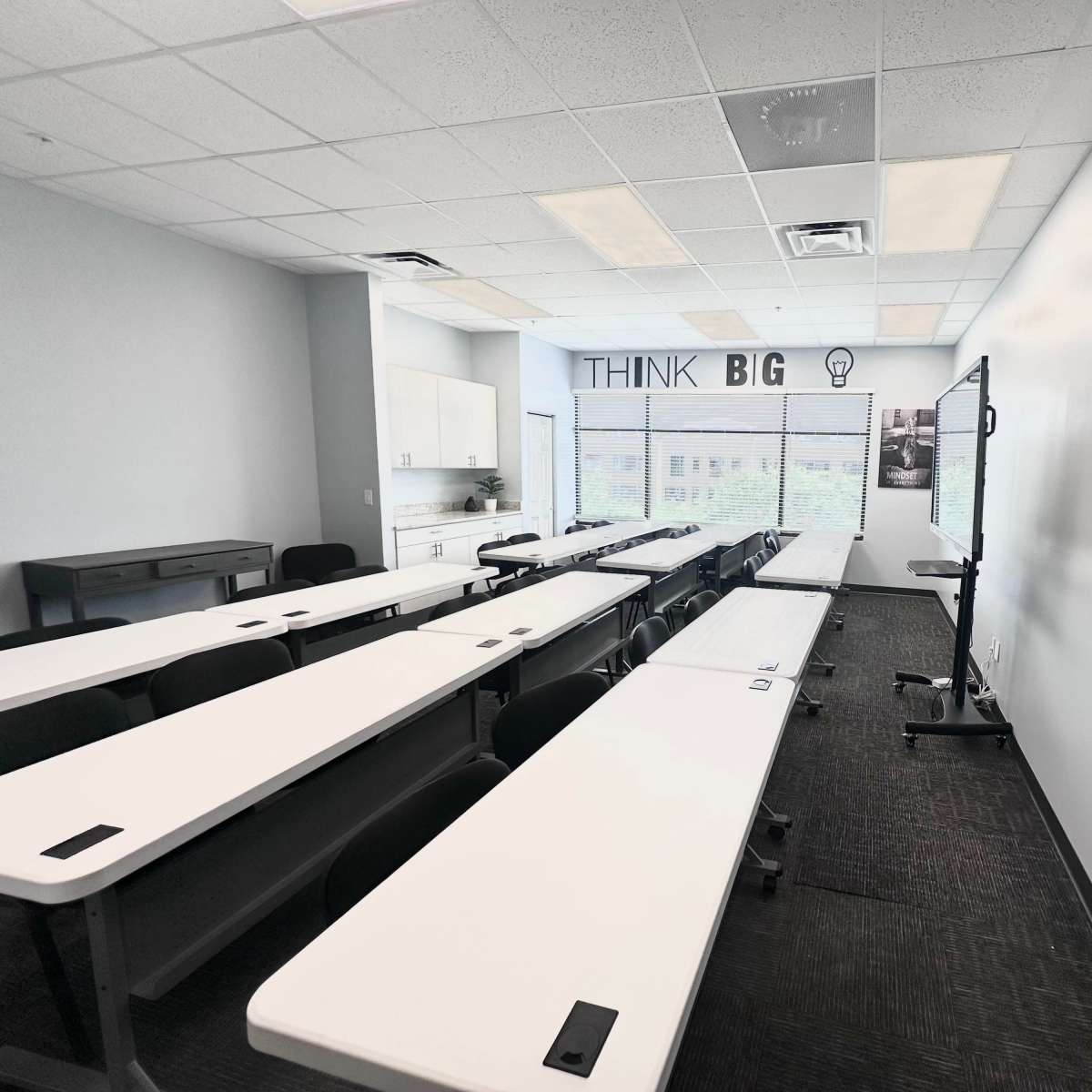 A modern classroom features rows of white tables and chairs, with a large screen and motivational wall art that says "THINK BIG."