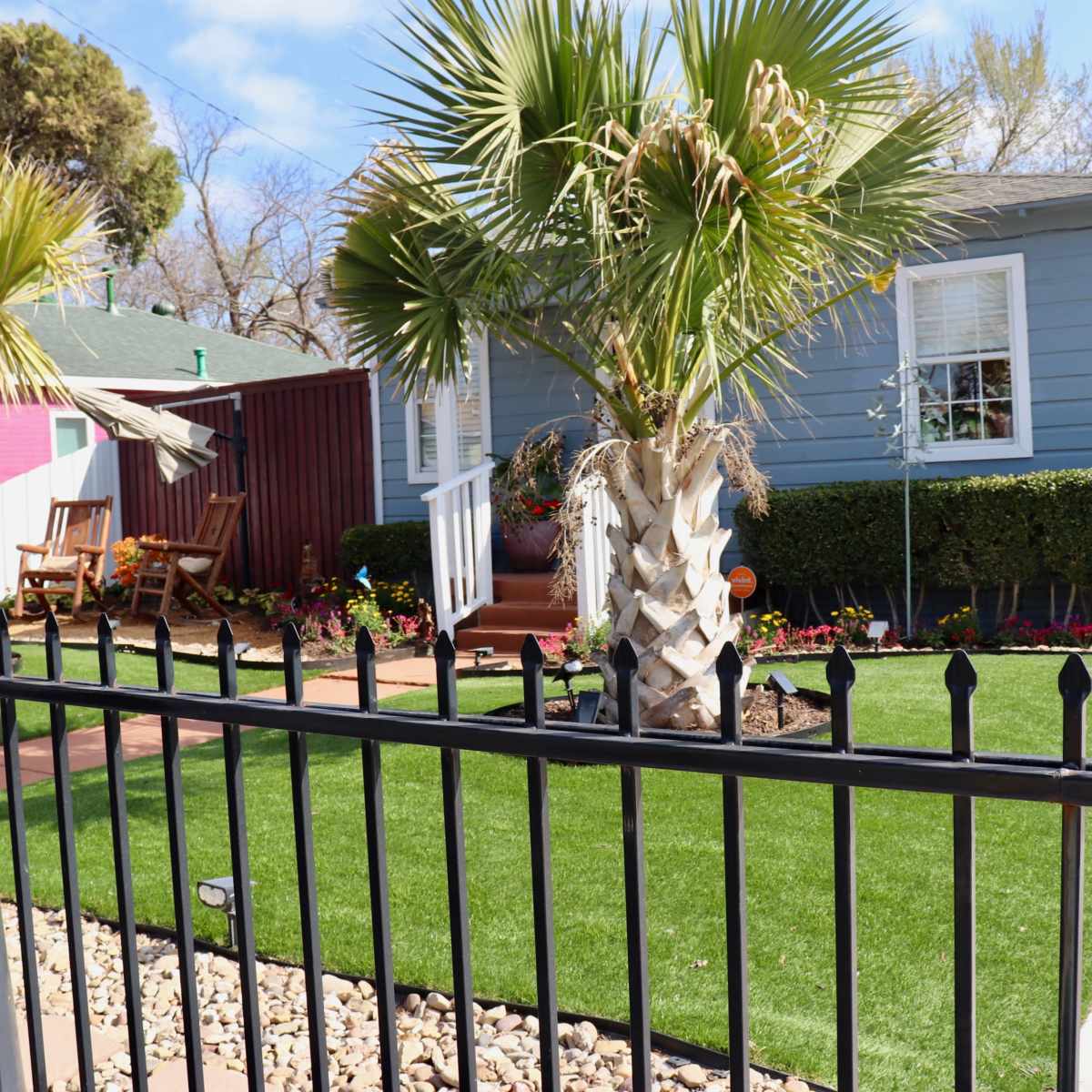 A house with a light blue exterior, surrounded by a well-maintained yard featuring palm trees, a green lawn, and a decorative fence.