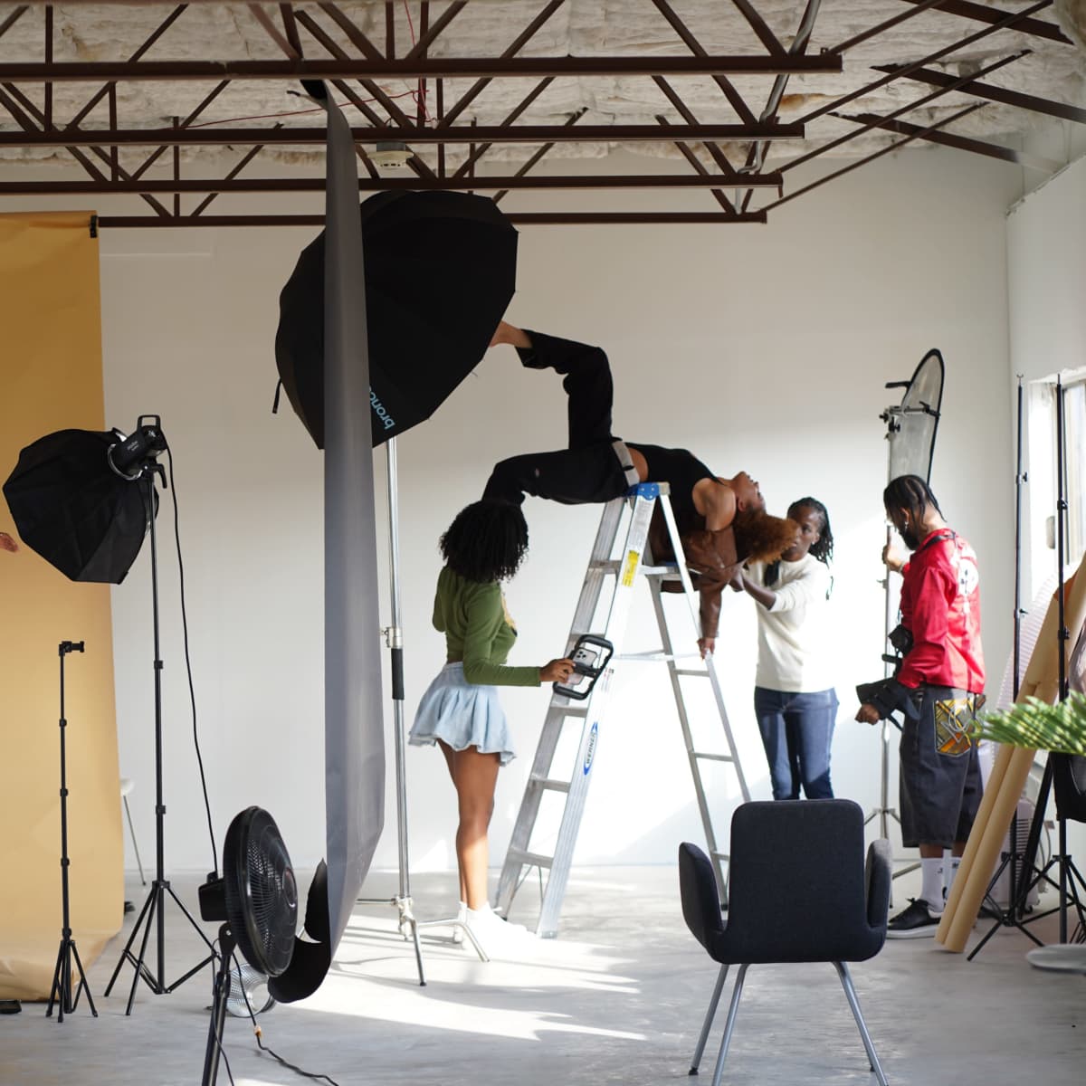 A photography studio features a model posing on a ladder while several photographers and assistants adjust lighting and equipment.
