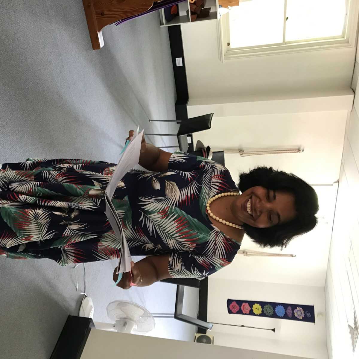 A woman in a floral dress stands barefoot in a room holding papers and smiling.