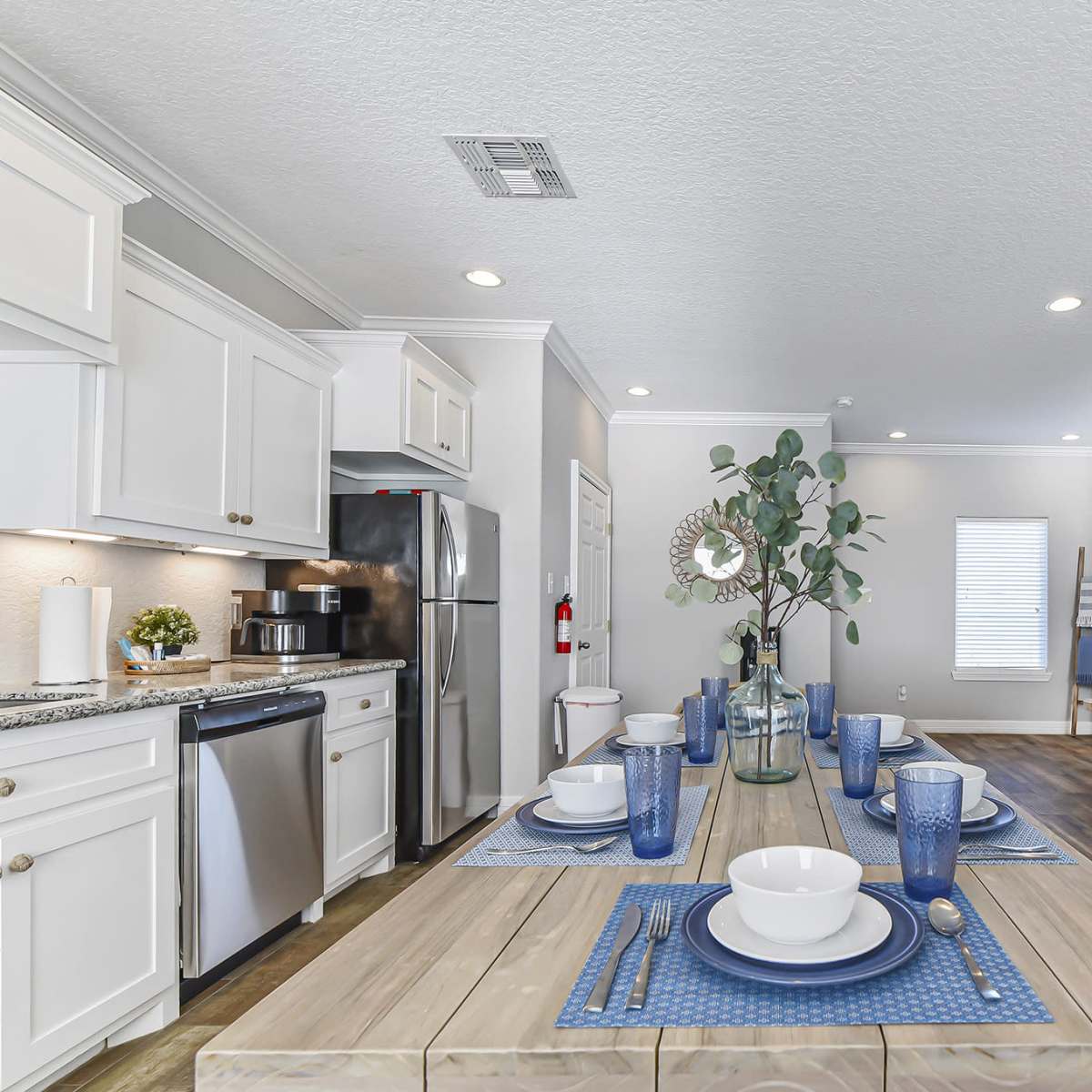 A well-lit modern dining area features a wooden table set for six, with blue and white tableware, adjacent to a contemporary kitchen and living area.