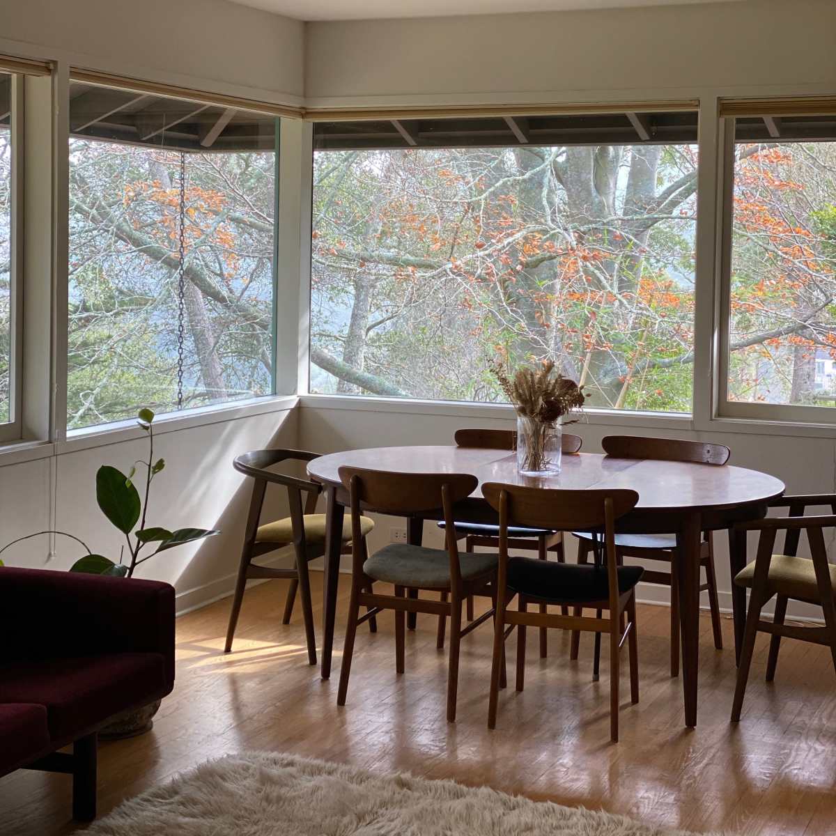 A dining area features a round table surrounded by wooden chairs, with large windows offering a view of trees outside.
