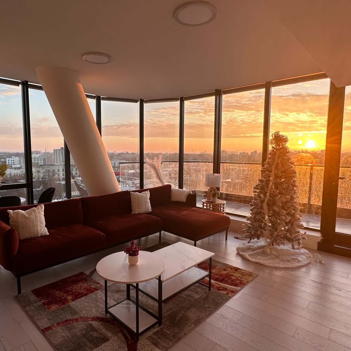 A modern living room features a sofa, coffee table, and a decorative Christmas tree, with large windows showcasing a sunset view over the city.