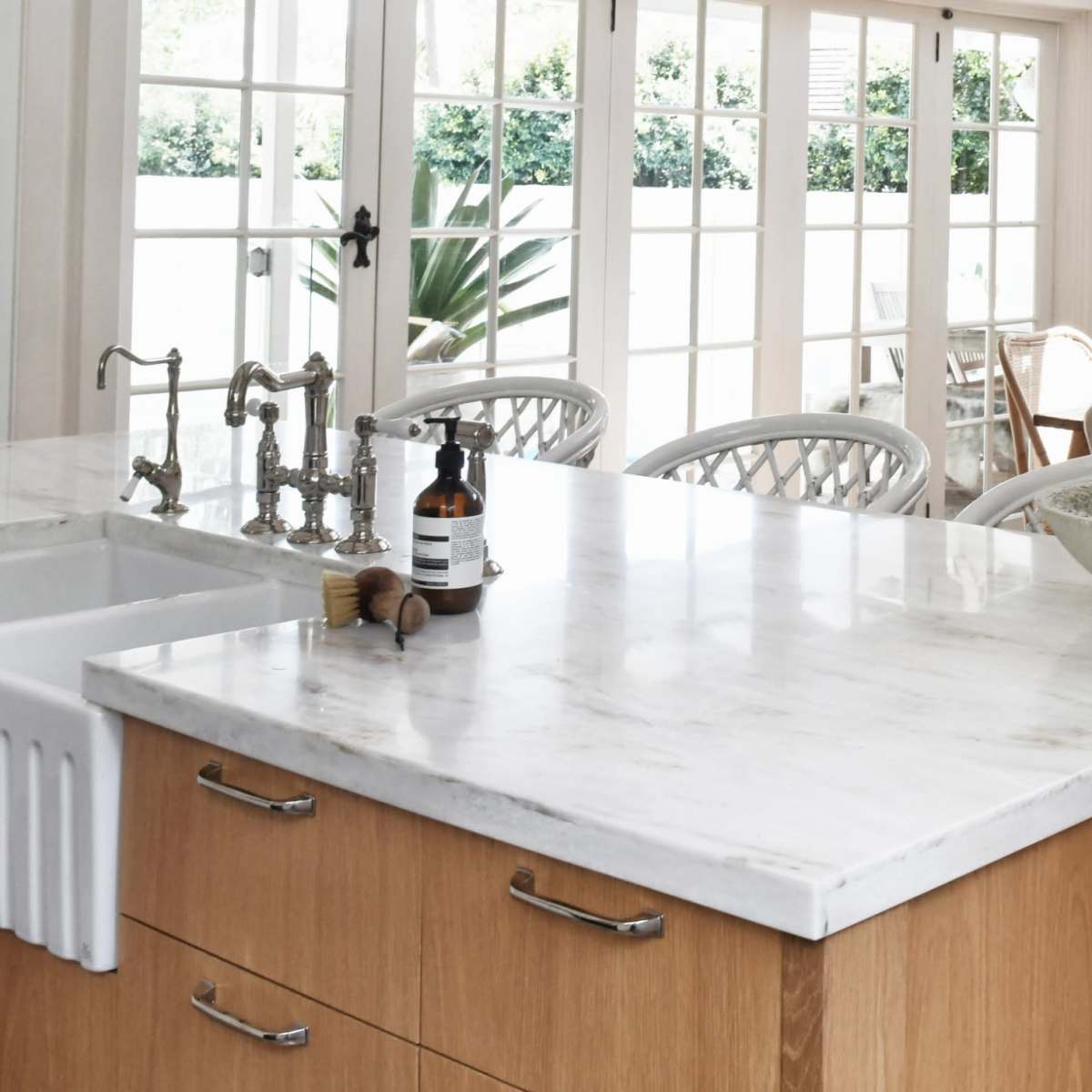 A modern kitchen with a marble countertop island and a dining area featuring a wooden table and chairs, illuminated by large windows.