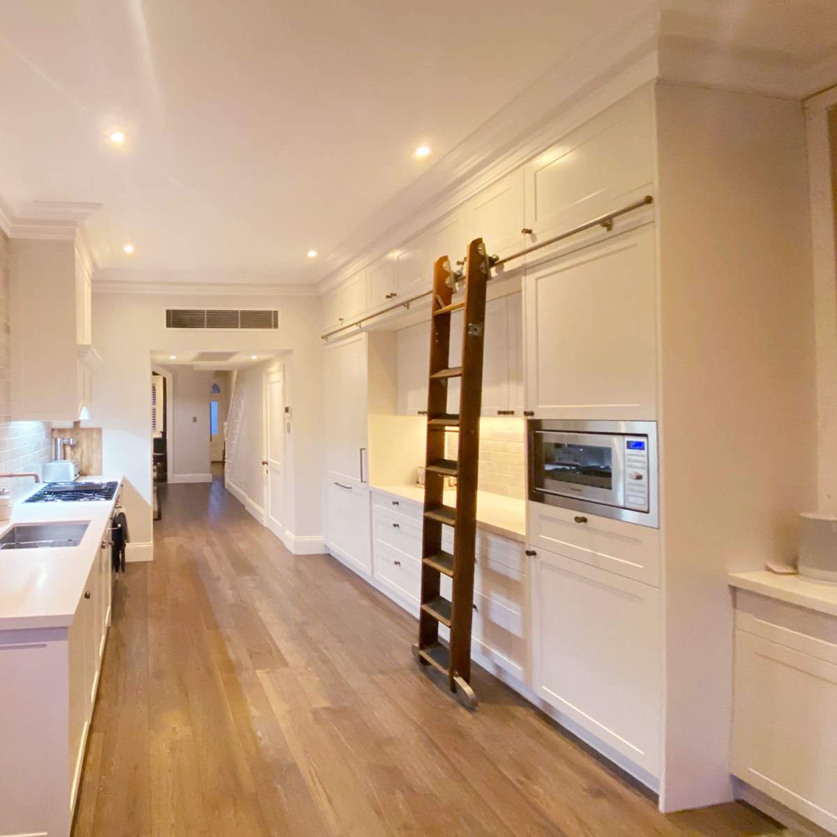 A modern kitchen with white cabinetry, a large window, and a wooden ladder against the wall.