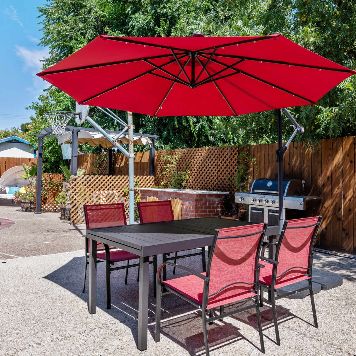 A patio area features a table with red chairs and a large red umbrella near a swimming pool and a barbecue grill.