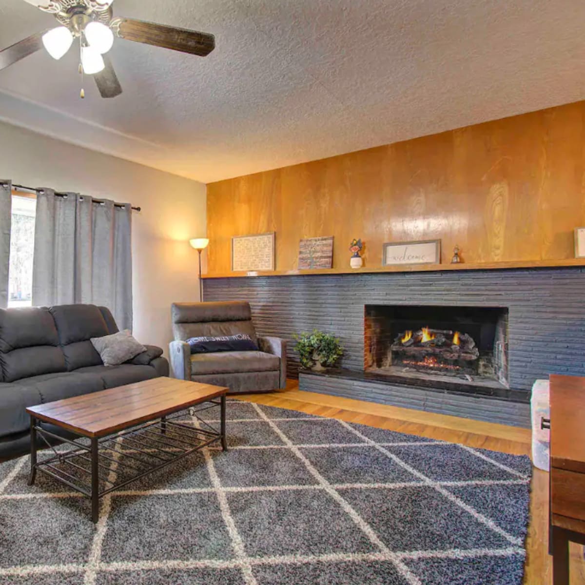 The living room features a fireplace, a wooden accent wall, and comfortable seating arranged around a central coffee table on a patterned rug.