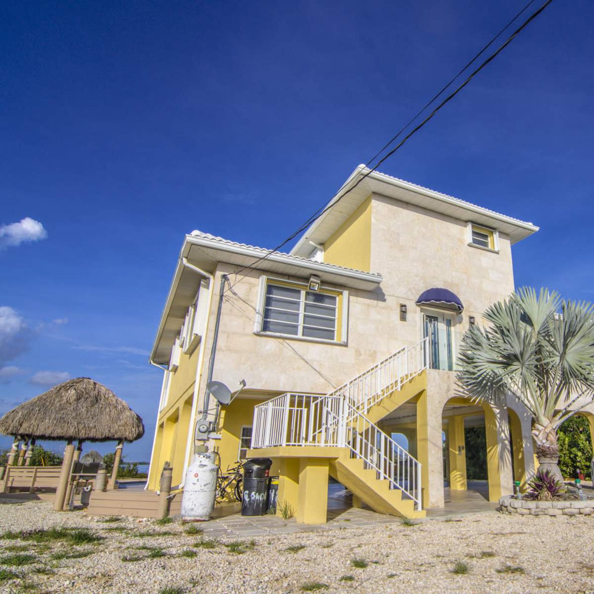 A two-story yellow house with a palm tree in front and a thatched roof structure nearby sits on a gravel driveway under a clear blue sky.