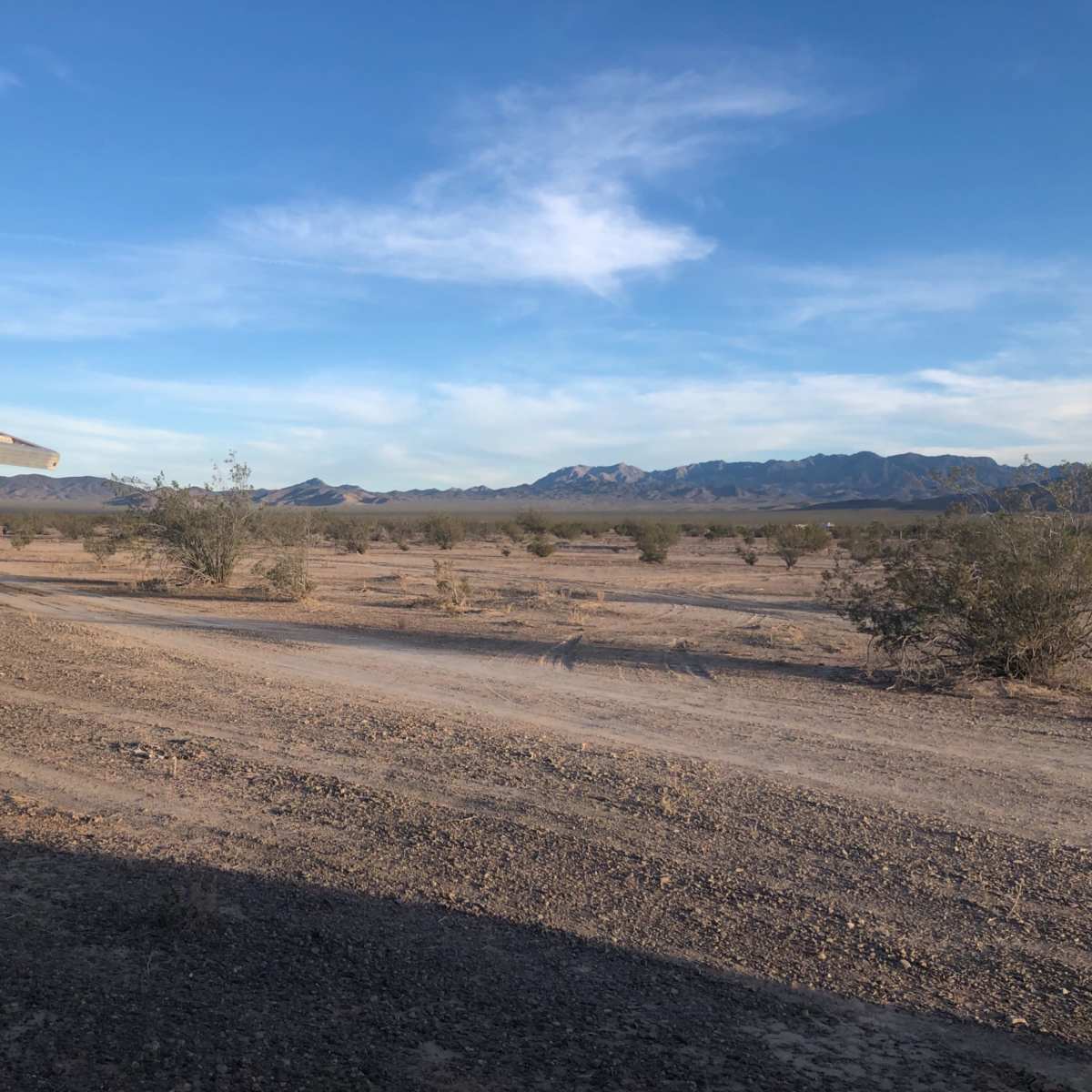 A rugged dirt road leads through a sparse desert landscape with distant mountains under a clear blue sky.