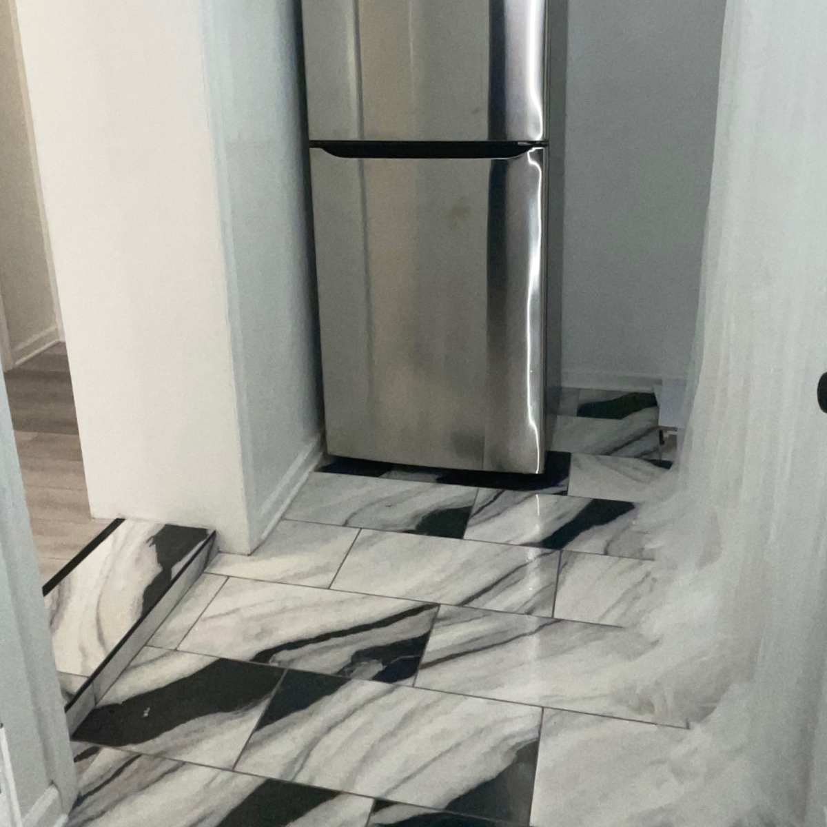 A stainless steel refrigerator is positioned against a wall on a floor with black and white marbled tiles.