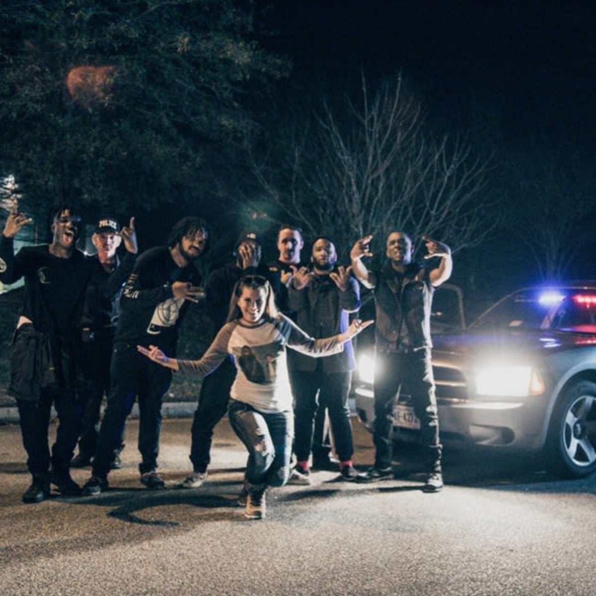 A group of eight people poses in front of a police car with its lights flashing on a dark street.