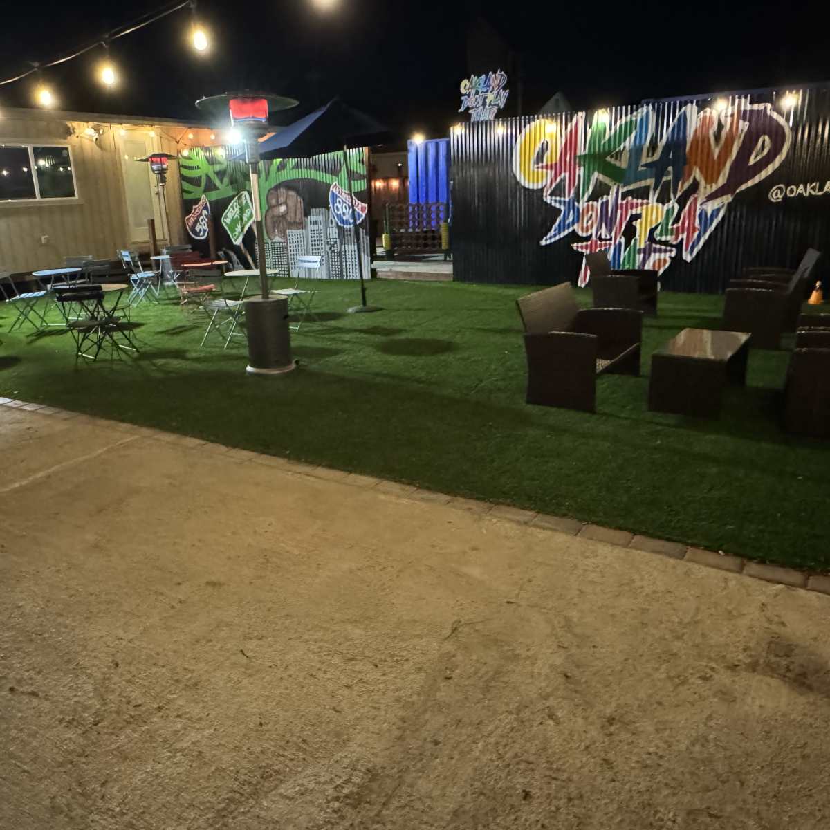 An outdoor seating area with string lights and decorative murals, featuring grass-like flooring and a mix of tables and chairs.