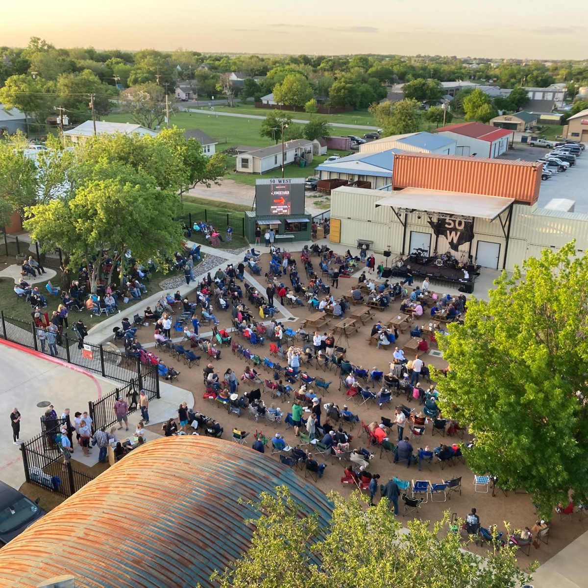 A large outdoor audience sits in folding chairs, arranged in rows, facing a stage at a community event area.