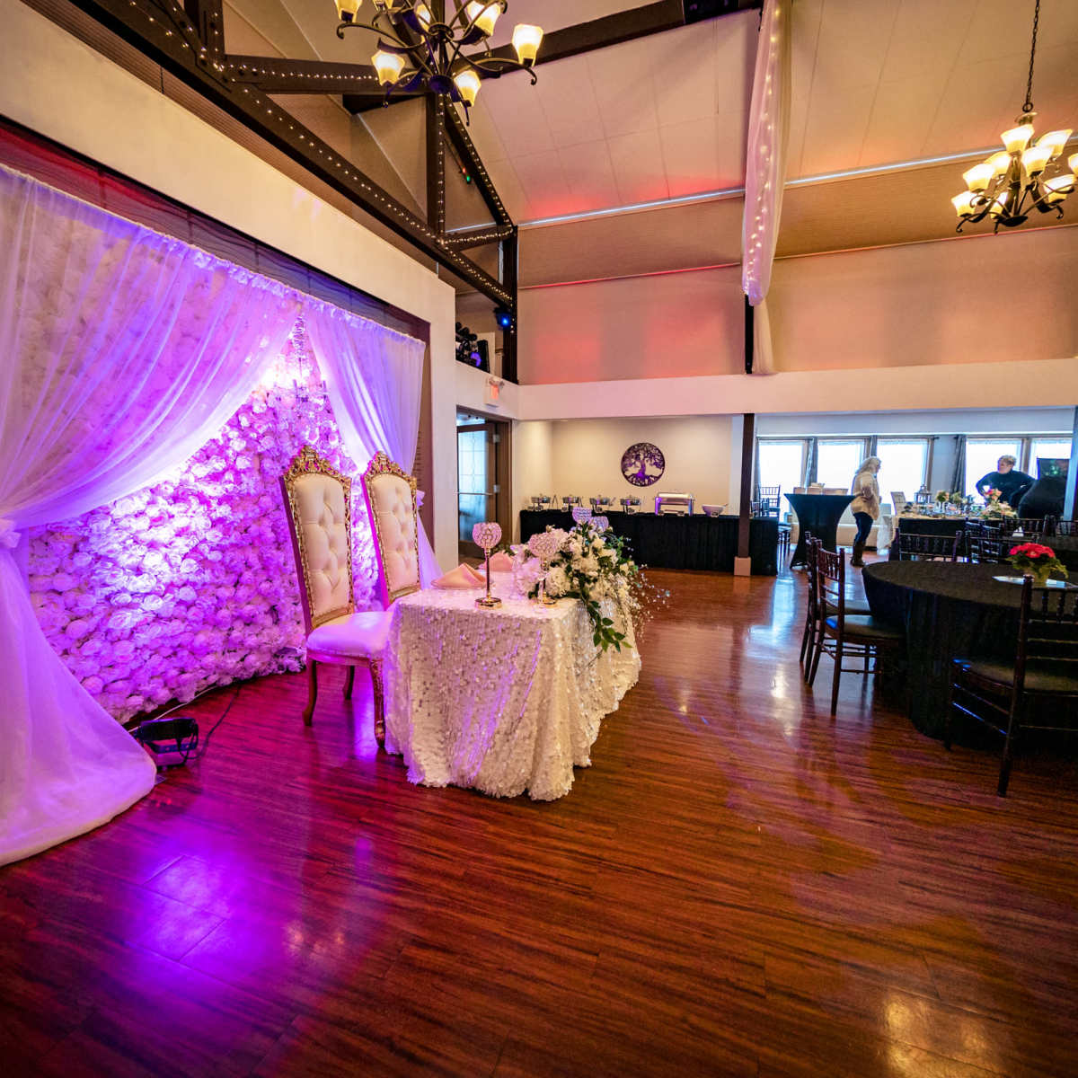 A decorated wedding reception area features a floral backdrop, a head table adorned with a white tablecloth, and dark wooden chairs in a spacious venue.