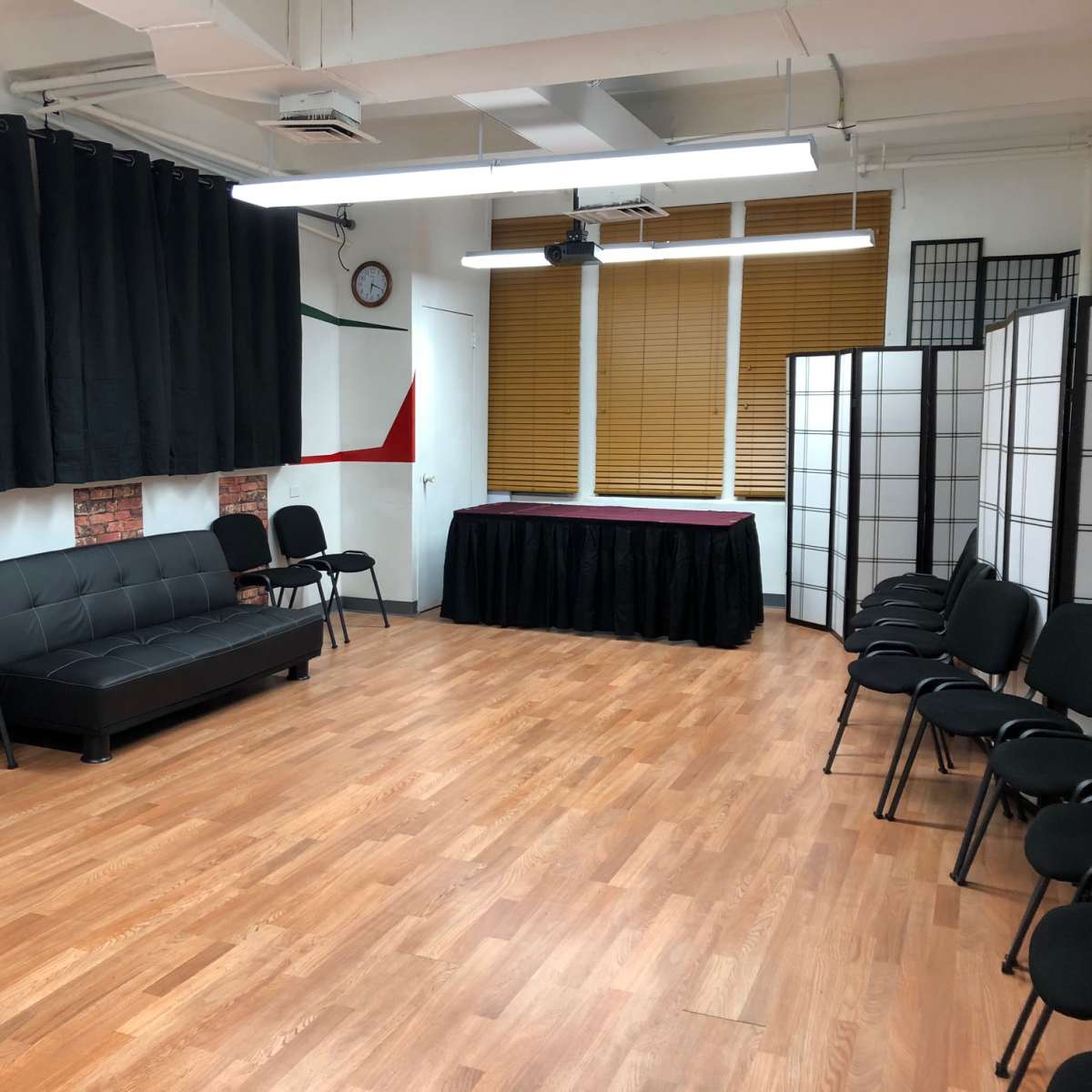 An empty room features a black sofa, several chairs arranged around the space, and a table draped in black fabric at the front.