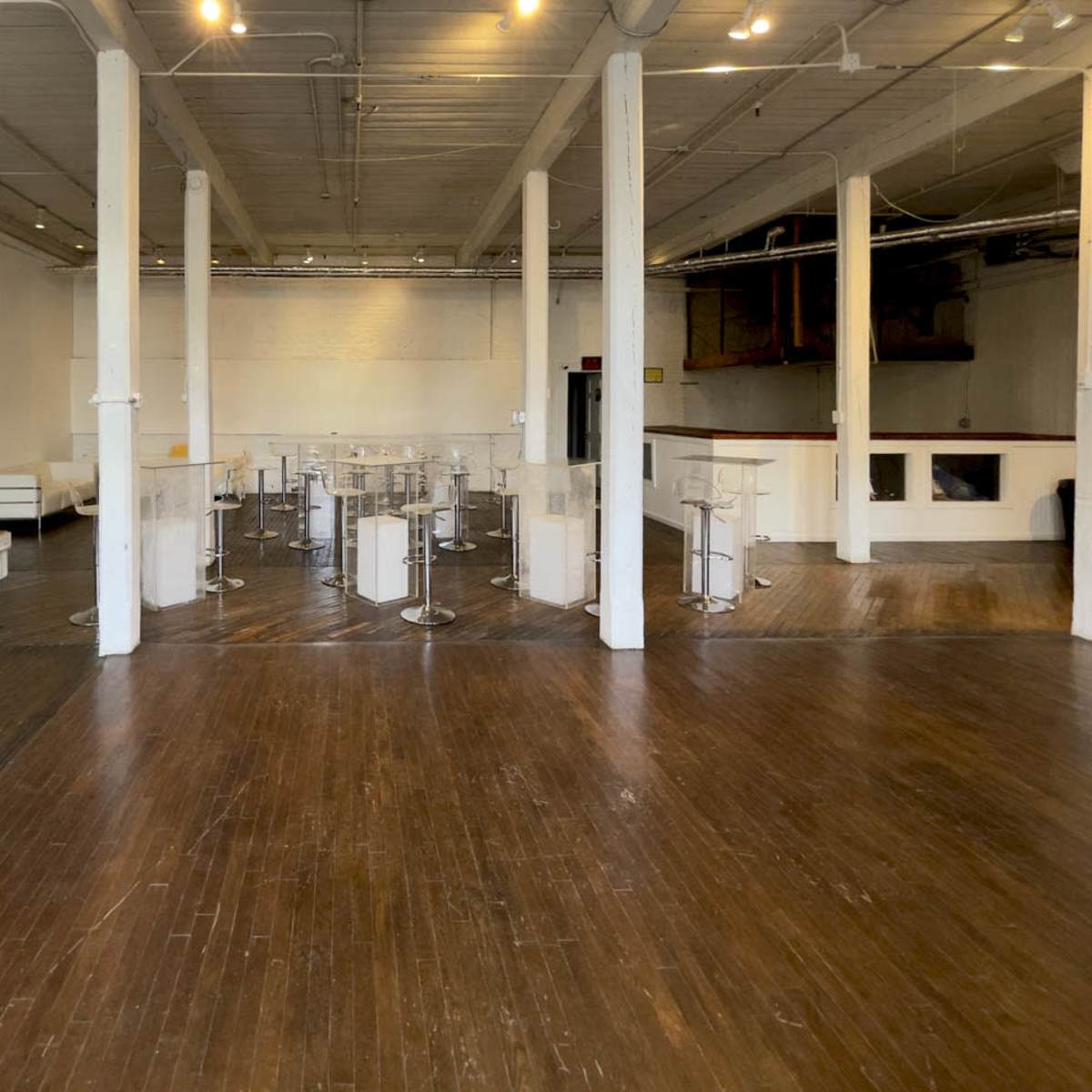 An empty indoor space with polished wooden floors, white walls, and several transparent bar stools arranged around small tables.
