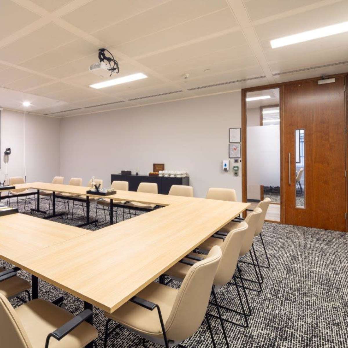 A conference room features a large U-shaped table surrounded by chairs, with a presentation screen and a wooden door in the background.