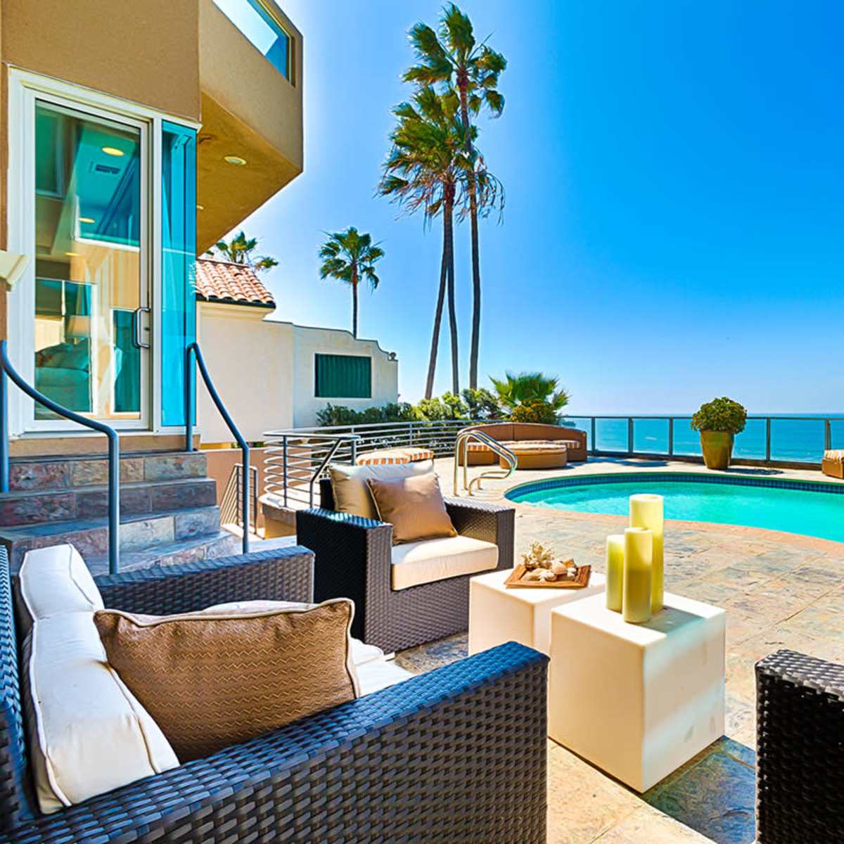 A patio area featuring wicker seating and a small table overlooks a swimming pool beside the ocean, framed by palm trees under a clear blue sky.