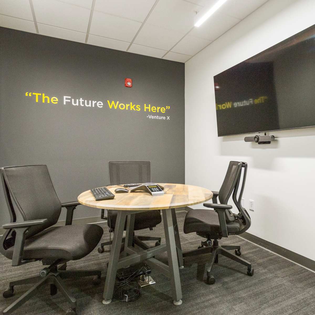 A small conference room features a round wooden table with three black chairs, a wall-mounted television, and a quote displayed on the wall.