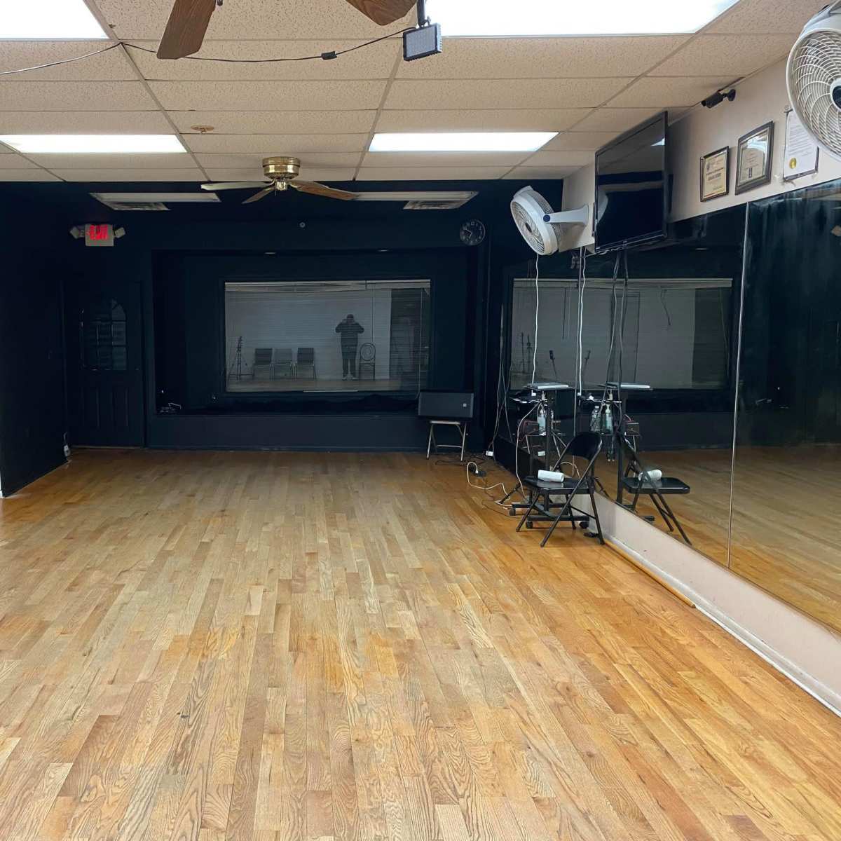 An empty dance studio with wooden flooring, black walls, a large mirror, and a ceiling fan.