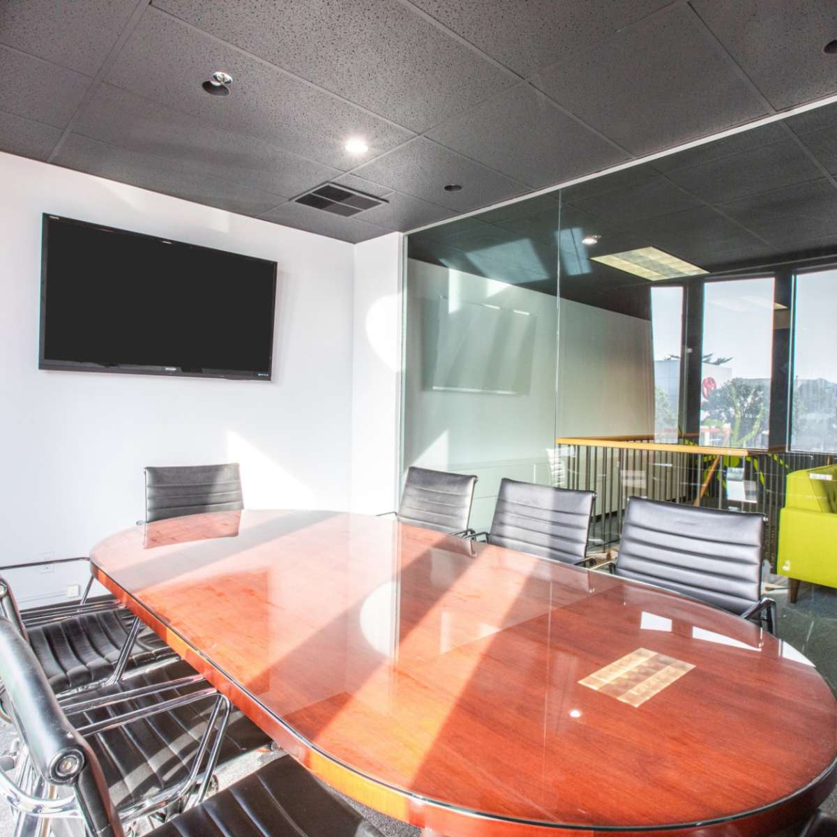 A modern conference room features a wooden oval table surrounded by black chairs, with a large glass wall and a television mounted on the wall.