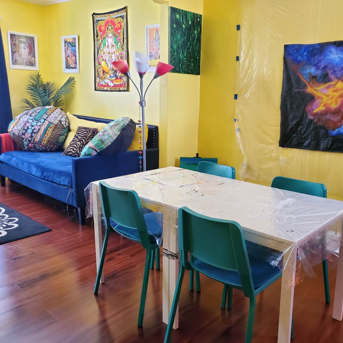 A bright yellow living room features a blue sofa with colorful cushions, a plastic-covered dining table with chairs, and various artworks on the walls.