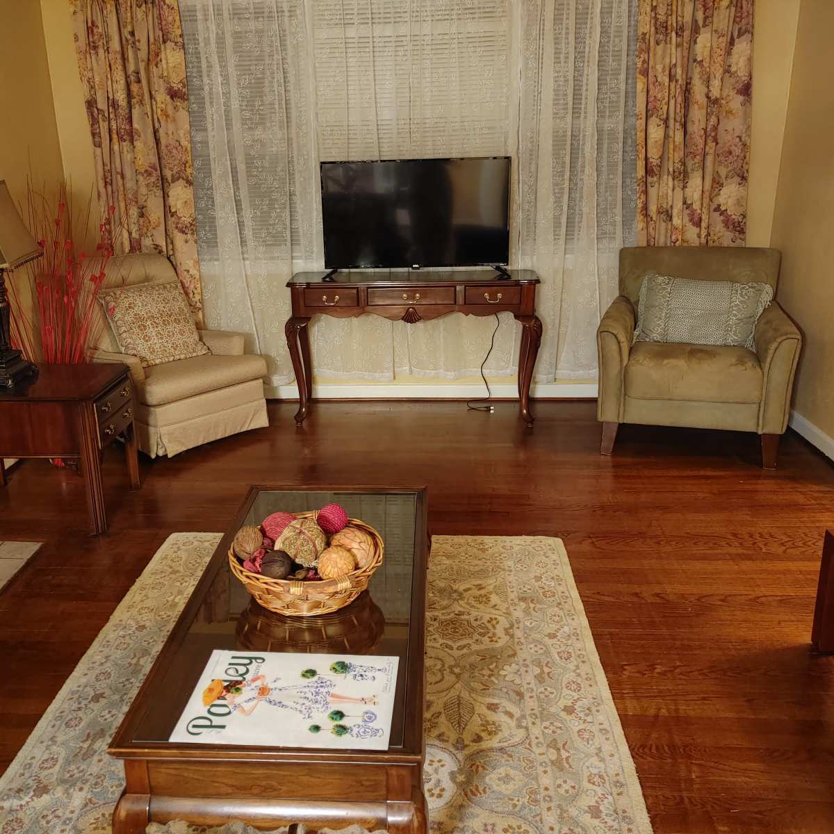 The living room features two chairs, a wooden coffee table with a decorative bowl, a TV stand with a flat-screen television, and a light-colored rug on polished wooden floors.