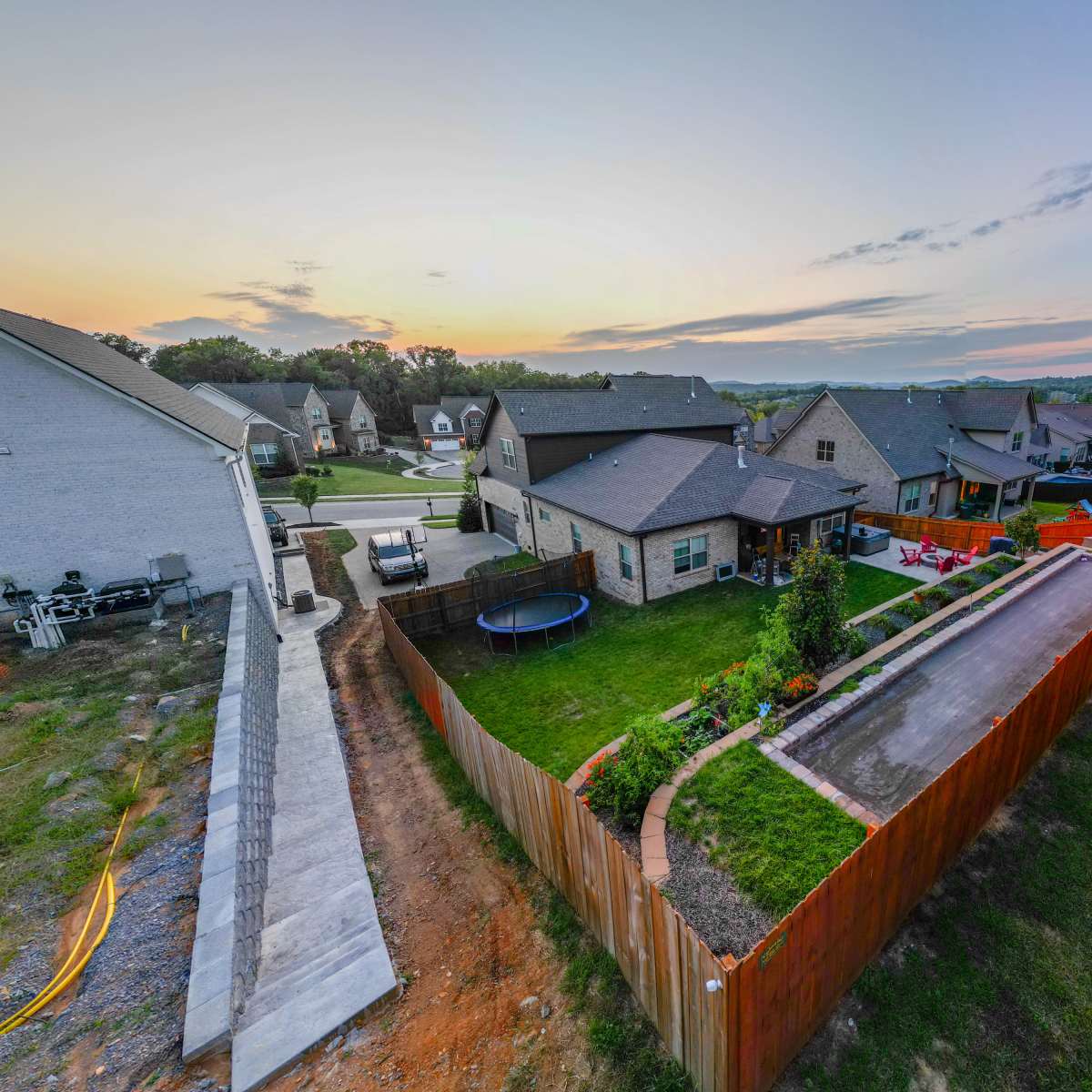 A suburban neighborhood with several houses, a backyard garden, a trampoline, and a view of a sunset in the background.