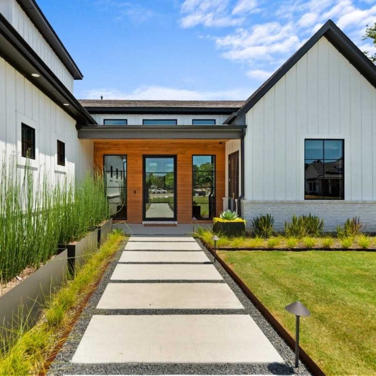 A modern home entryway with a stone walkway leading to large wooden double doors, bordered by neatly arranged grasses and shrubs.