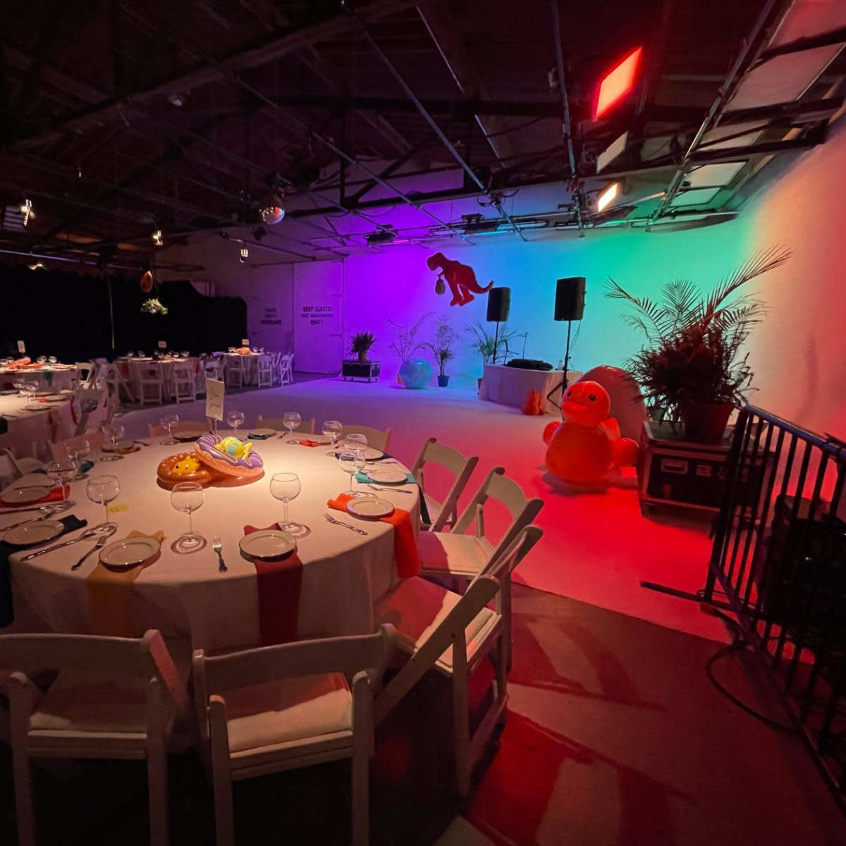 A decorated event space with round tables set for dining, colorful lighting, and stage elements in the background.