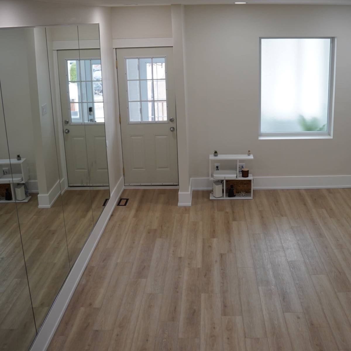 A spacious, well-lit room with light wooden flooring, a large mirror on one wall, a door, and minimal furnishings, including a small table and a decorative plant.