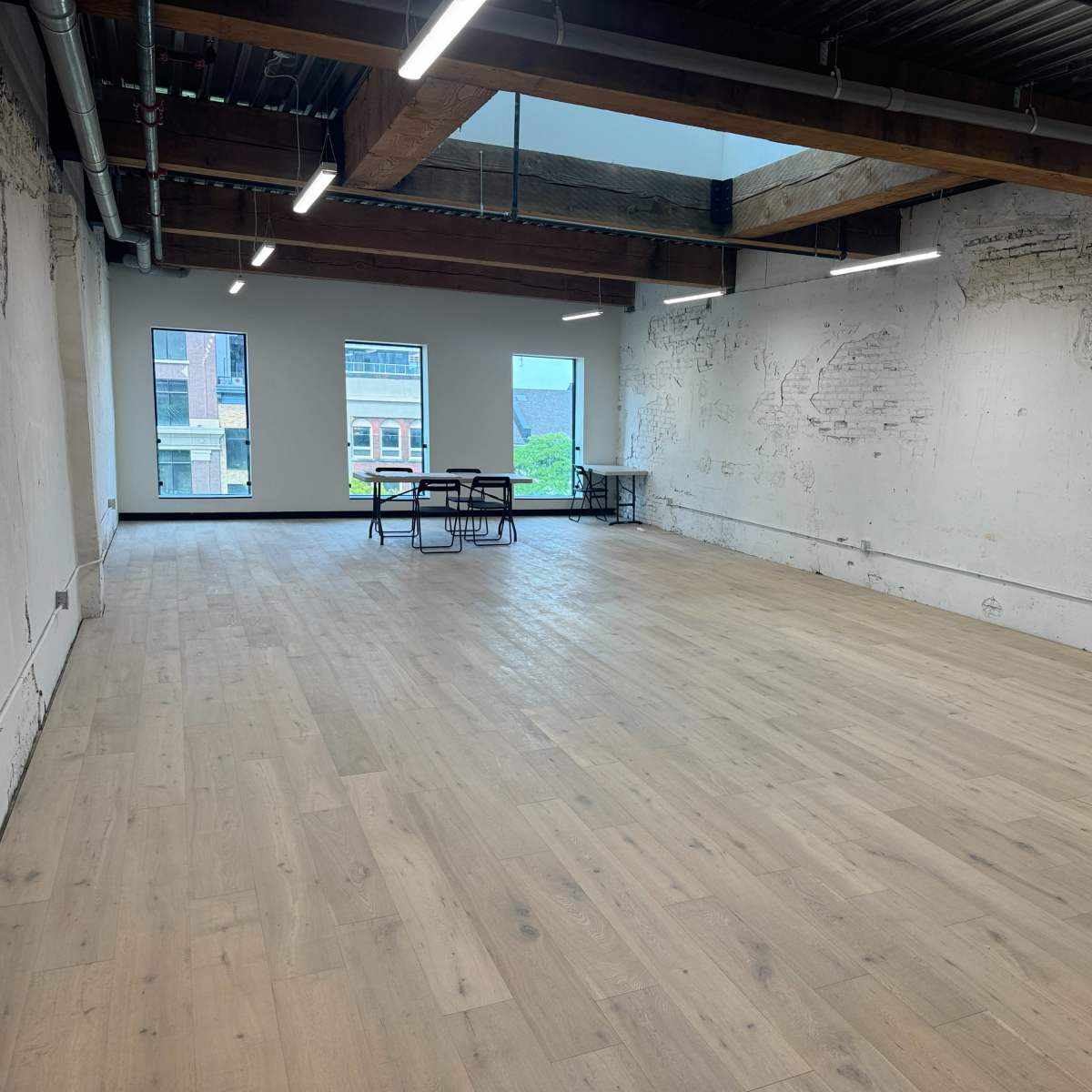 A spacious, unfinished interior room features large windows and wooden beams overhead, with a few tables positioned in the center and bare walls.