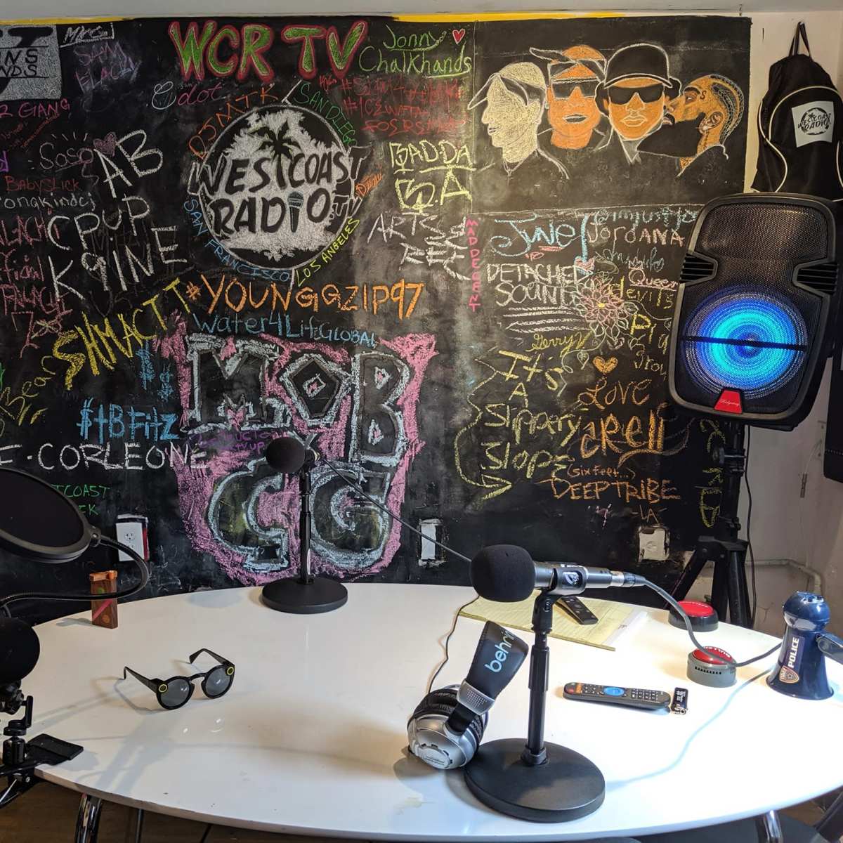 A radio studio with a circular table surrounded by microphones, a colorful spinning wheel, and a chalkboard wall covered in colorful writings and drawings.