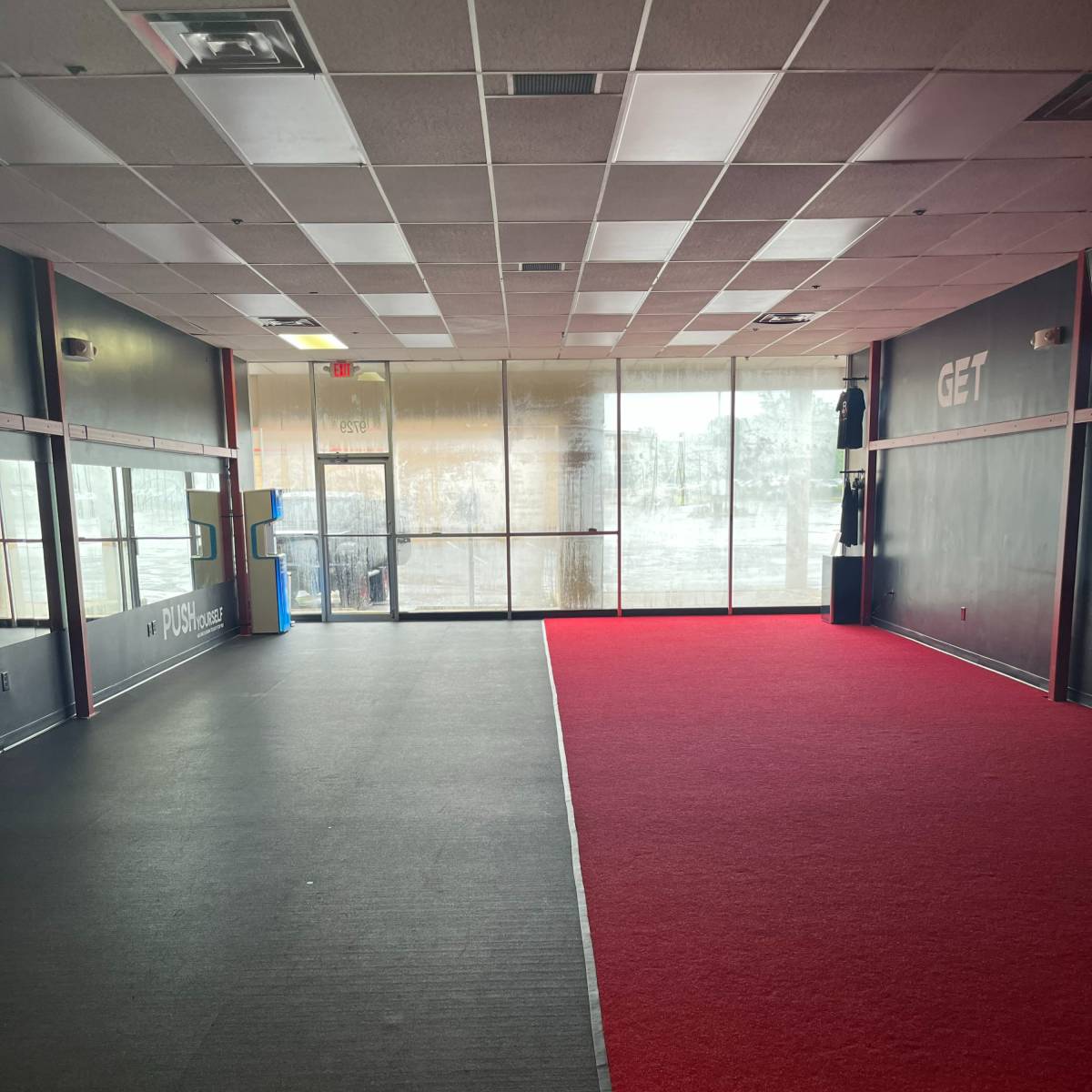 A large, empty room with black rubber flooring on one side and red carpet on the other, featuring large windows along one wall.