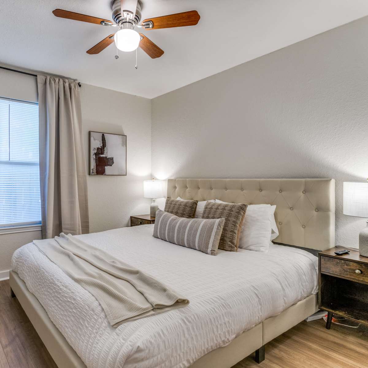 A neatly arranged bedroom features a large bed with neutral bedding, side tables with lamps, and a window beside a potted plant.
