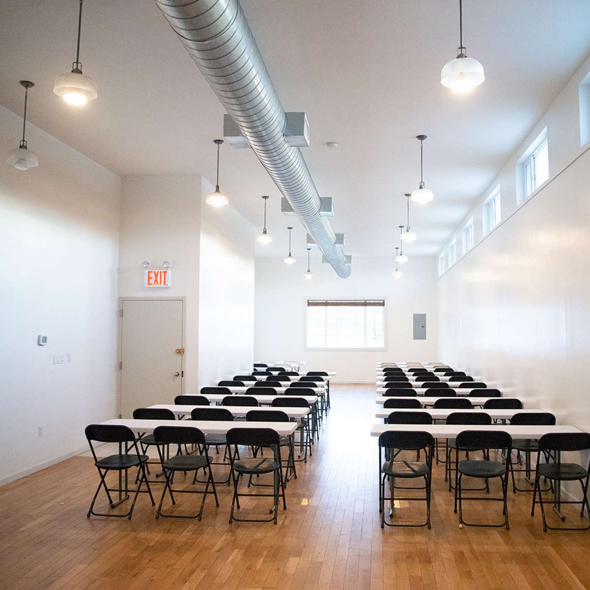 A row of black folding chairs is arranged in a rectangular formation in a bright, sparsely decorated room with large windows and overhead lights.