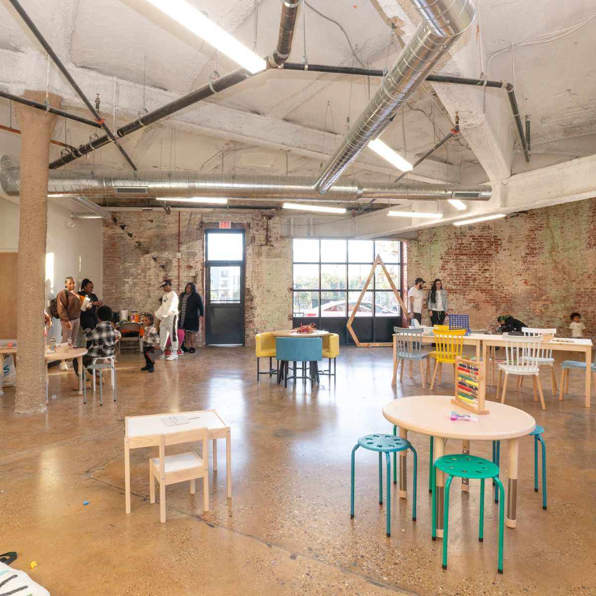 A spacious, modern classroom features several tables and chairs, with children and adults engaging in activities near large windows and exposed brick walls.