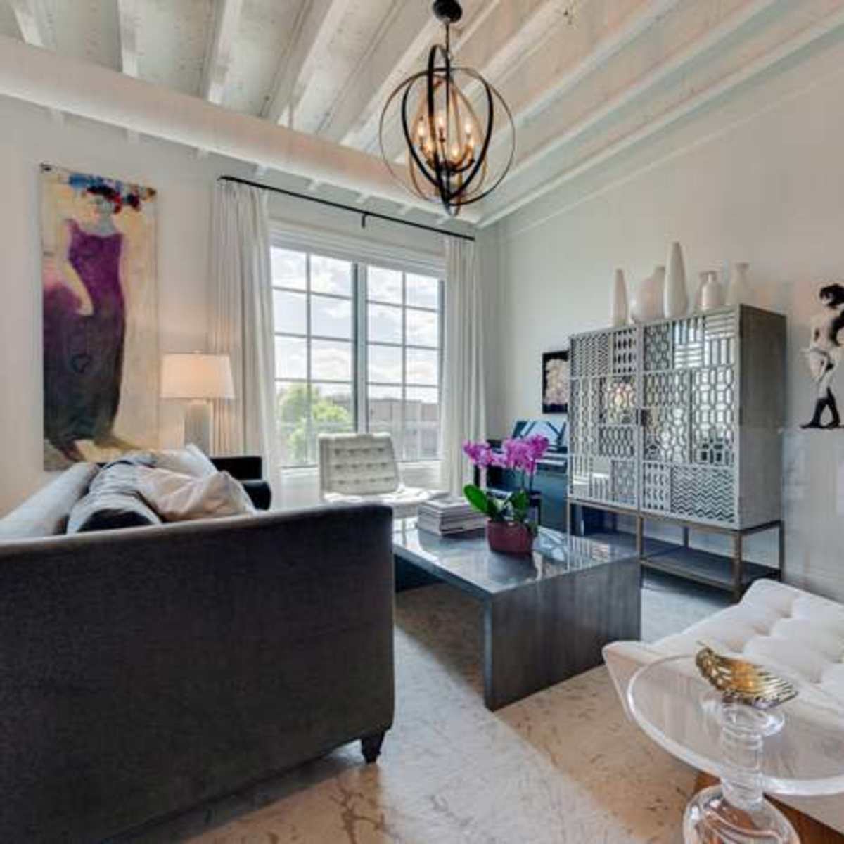The room features a gray sofa, a glass coffee table, and a white chair, with large windows allowing natural light to enter.