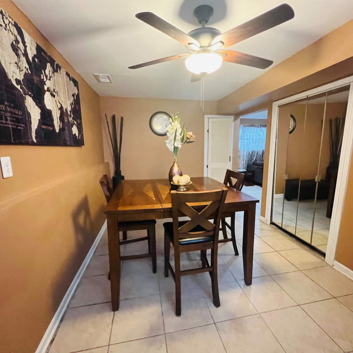A dining area features a wooden table with four chairs, a ceiling fan, and a wall adorned with a world map, alongside a mirrored closet.