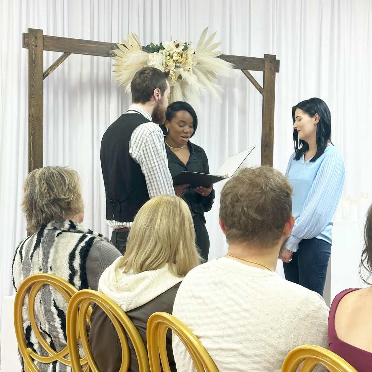 A couple stands in front of a person reading from a booklet during a ceremony, with guests seated on gold chairs in the foreground.