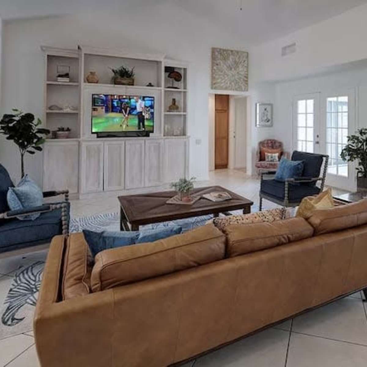 The living room features a leather sofa, two blue chairs, a wooden coffee table, and a television mounted on a shelf against a light-colored wall.