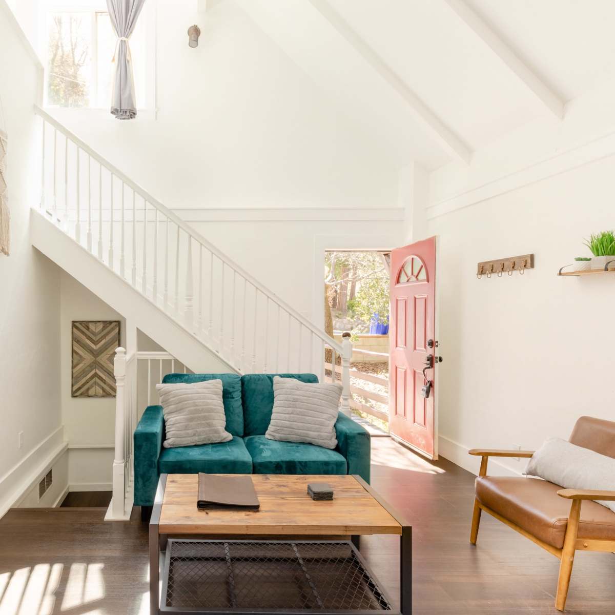 A bright interior with a teal sofa, wooden coffee table, and a staircase leading to an upper level, with a pink door open to a view outside.