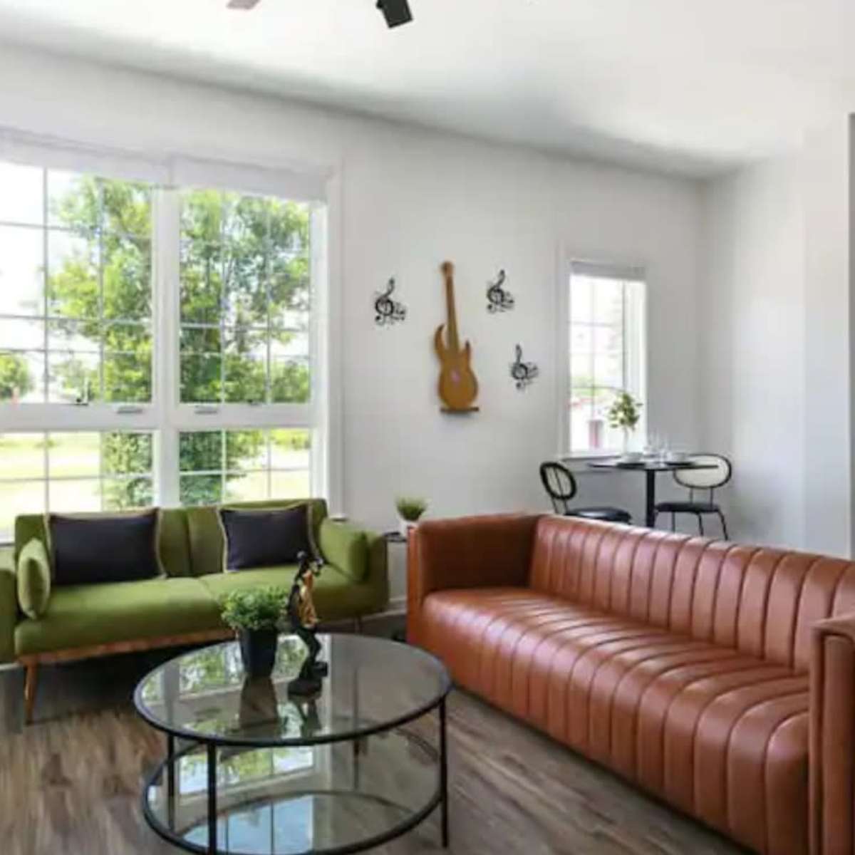 The living room features a green sofa and a brown leather couch arranged around a round glass coffee table, with large windows allowing light to enter the space.