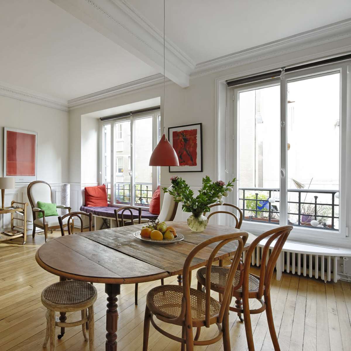 A bright living room features a round wooden table surrounded by wooden chairs, with a mix of artwork on the walls and plants by the windows.