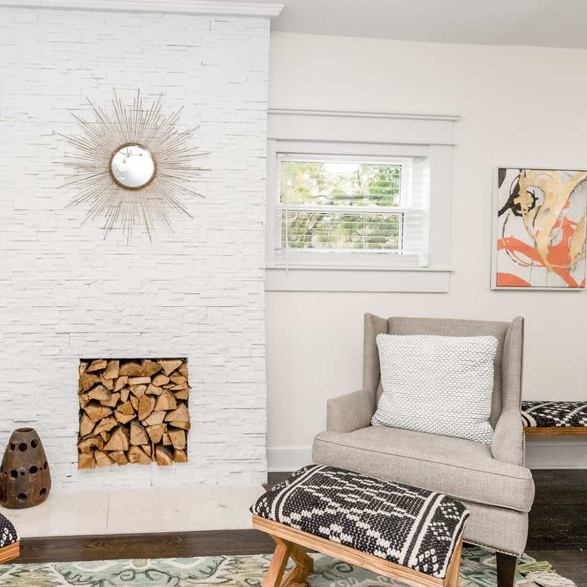 A cozy living room featuring two armchairs with patterned cushions, a stone fireplace with stacked wood, and decorative elements including a wall mirror and artwork.