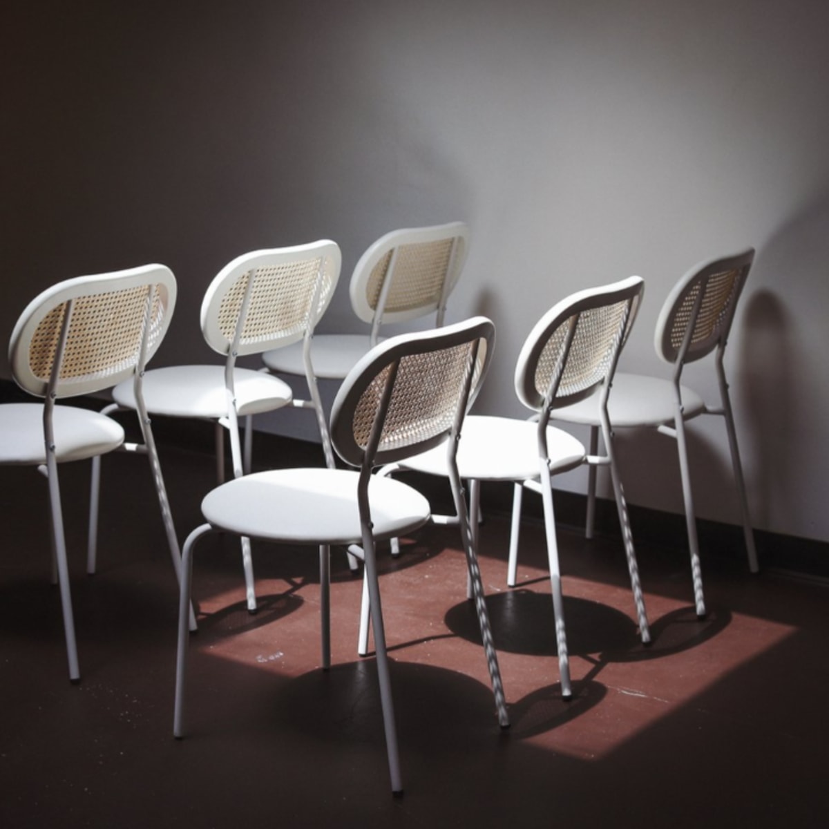 A row of white chairs with woven backs is arranged in a dimly lit room.