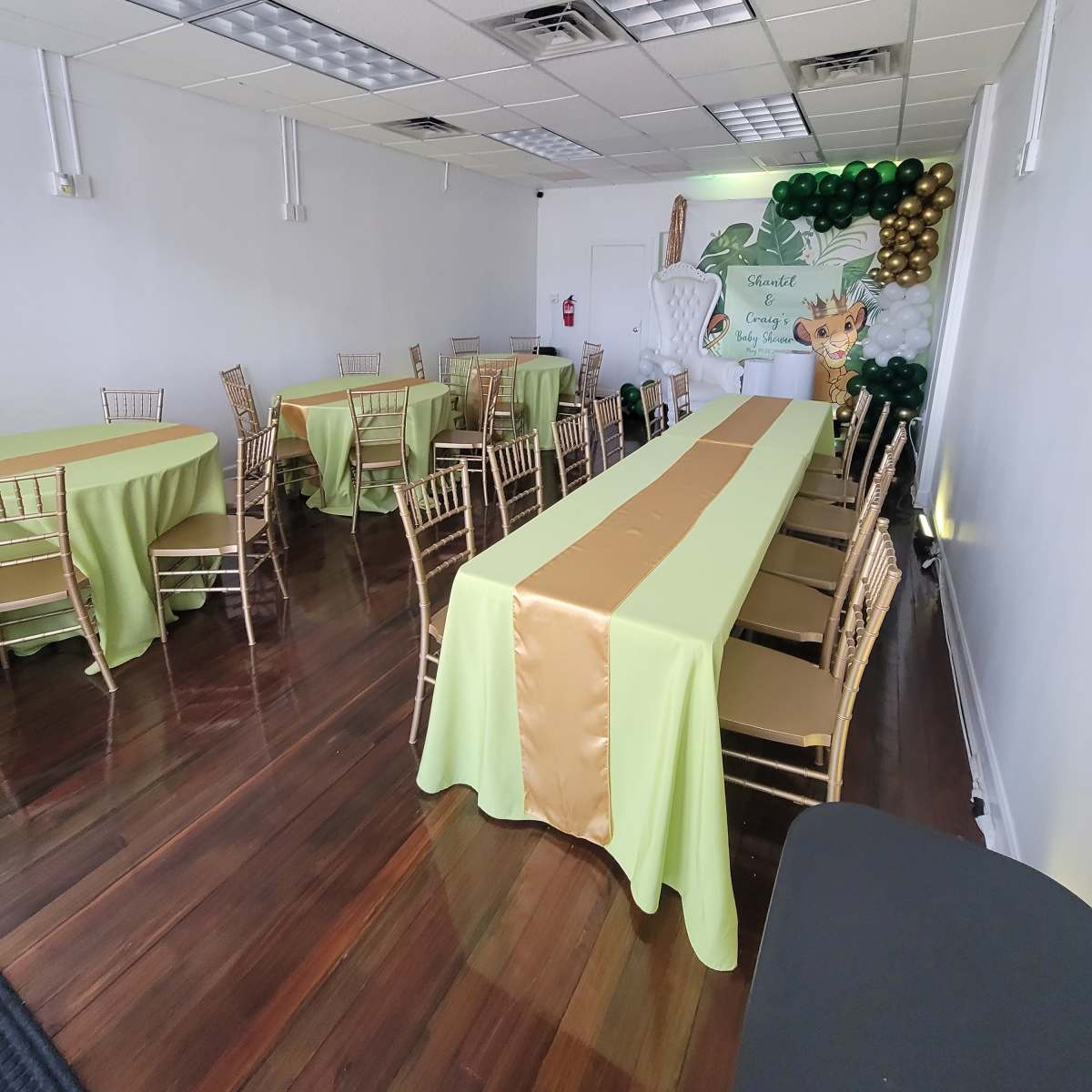 A brightly lit event space features several tables covered in green and gold linens, with decorative balloons and a backdrop on one wall.